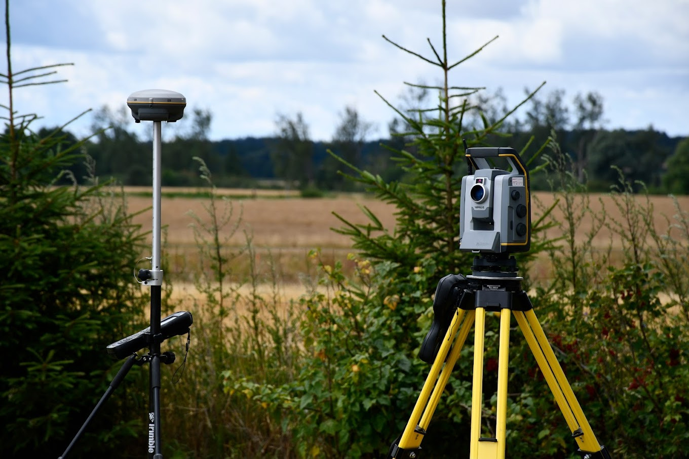Dwa urządzenia pomiarowe: odbiornik GNSS na statywie i tachimetr z żółtymi nogami na tle pola i drzew, widoczne krzewy i małe drzewka iglaste.
