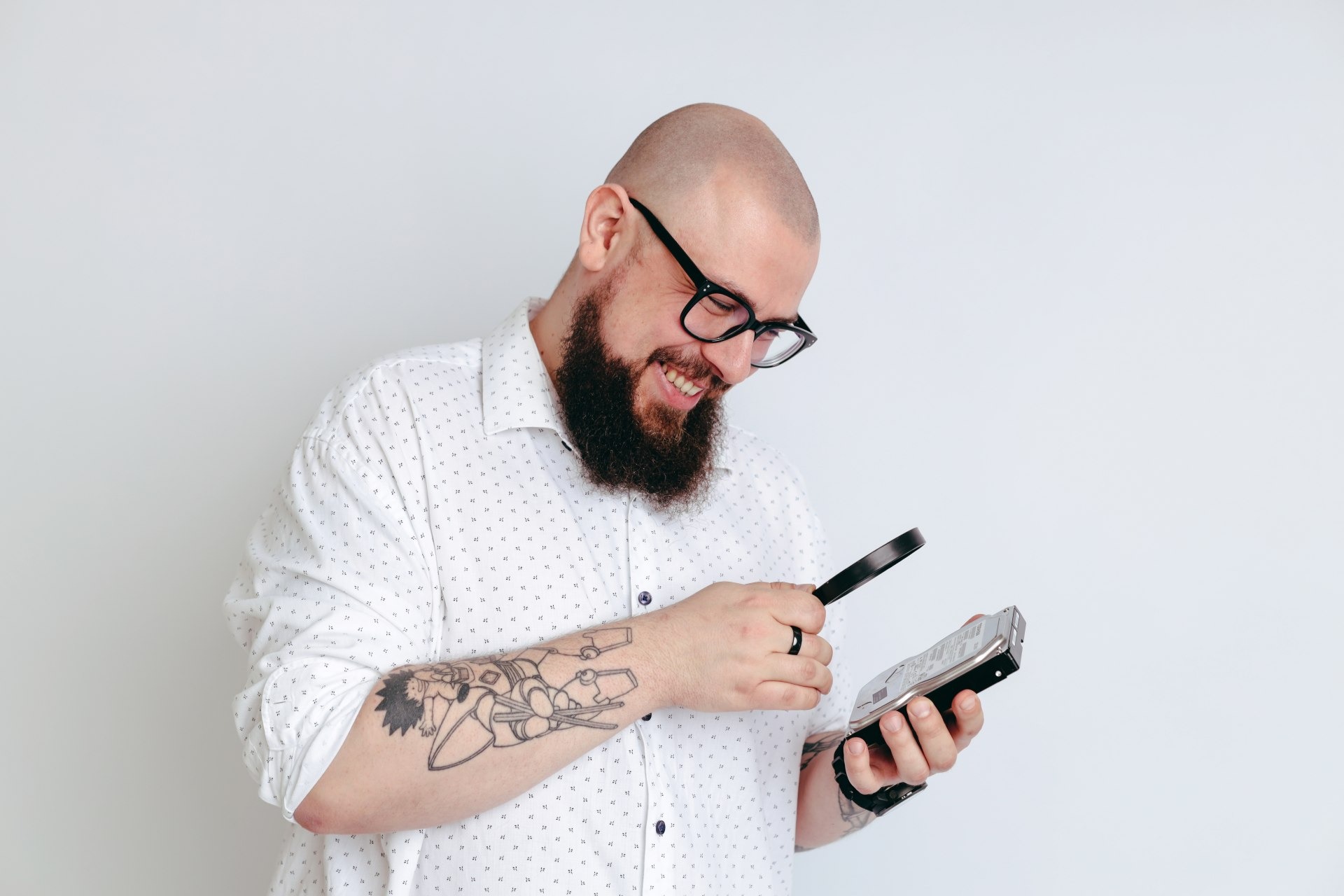 Uśmiechnięty mężczyzna z brodą i tatuażem na ramieniu, w okularach, analizuje dysk twardy przez lupę na białym tle.