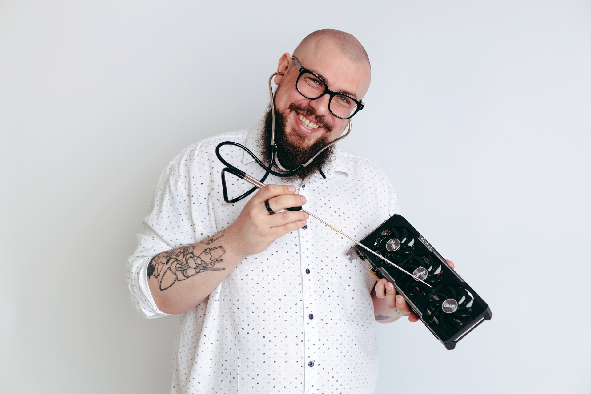 Uśmiechnięty mężczyzna z brodą i okularami, w koszuli w drobny wzór, używa stetoskopu i narzędzia do diagnostyki karty graficznej Sapphire Nitro na białym tle.