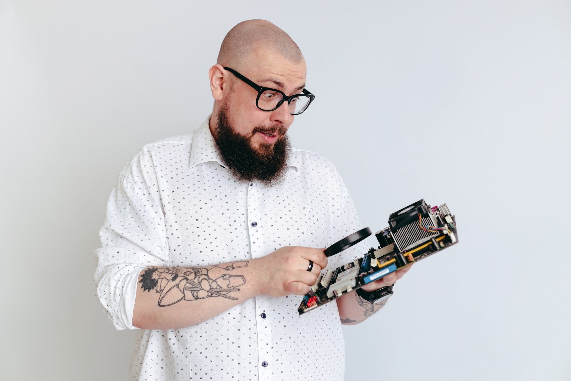 Mężczyzna z brodą i okularami, w koszuli w drobne wzory, z tatuażami na rękach, uważnie przygląda się płycie głównej komputera przez lupę na jasnym tle.