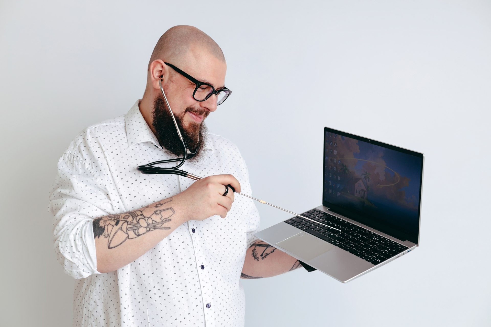 Łysy mężczyzna z brodą i tatuażami na ramieniu w okularach i białej koszuli w kropki, uśmiechnięty, bada stetoskopem otwartego laptopa na białym tle.