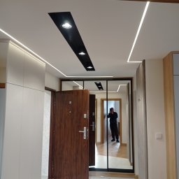 Danbud - Nowoczesny przedpokój z oświetleniem LED na suficie, brązowe drzwi wejściowe otwarte, duże lustro z czarną ramą, odbicie fotografa.