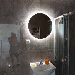 Danbud - Nowoczesna łazienka z umywalką, okrągłym lustrem z podświetleniem LED i kabiną prysznicową, wykończona płytkami imitującymi marmur, z widocznym odbiciem osoby robiącej zdjęcie.