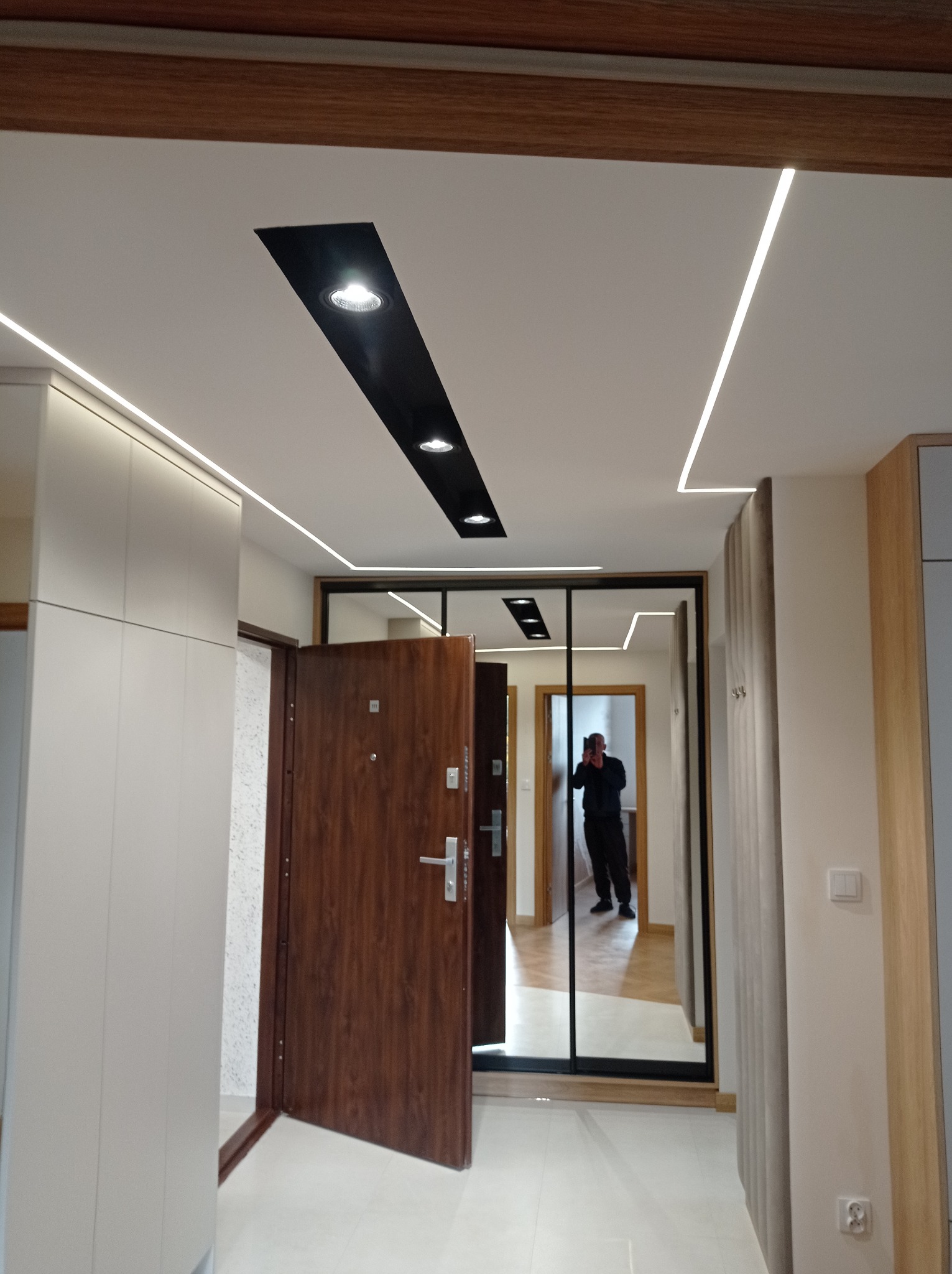 Nowoczesny przedpokój z oświetleniem LED na suficie, brązowe drzwi wejściowe otwarte, duże lustro z czarną ramą, odbicie fotografa.