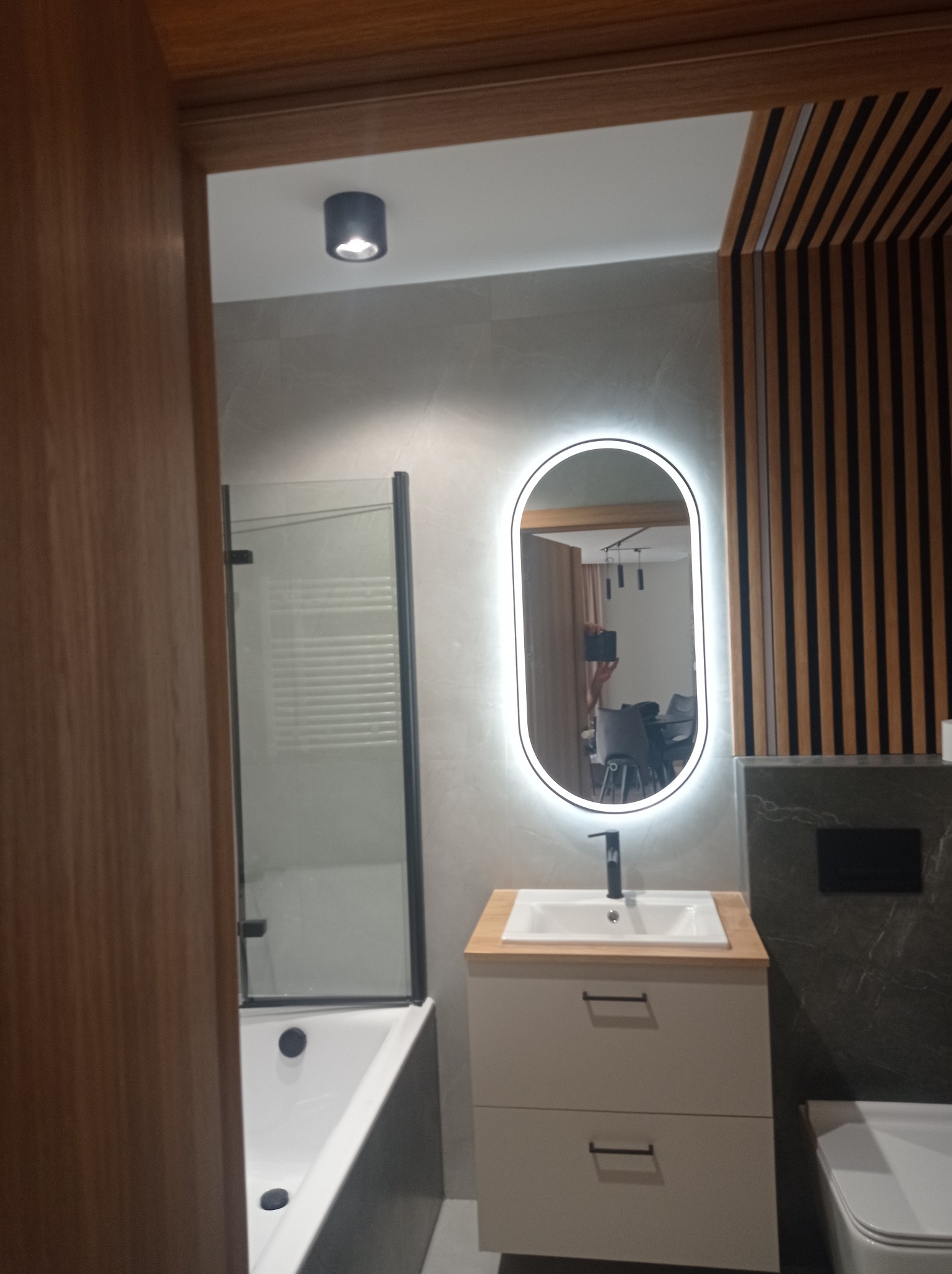 Nowoczesna łazienka z owalnym lustrem z podświetleniem LED, umywalką na szafce z drewnianym blatem, kabiną prysznicową z czarnymi ramami i dekoracyjną ścianą z drewnianych listew.