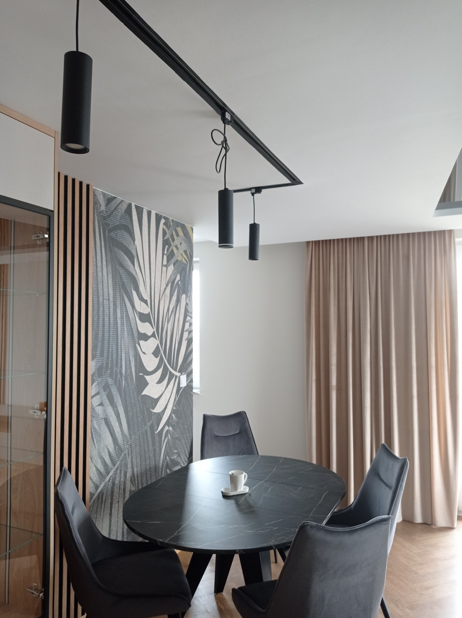 Jadalnia z okrągłym, czarnym stołem z marmurowym wzorem, otoczonym przez cztery szare krzesła; na stole biała filiżanka. W tle dekoracyjna ściana z motywem liści i zasłony w odcieniach beżu.