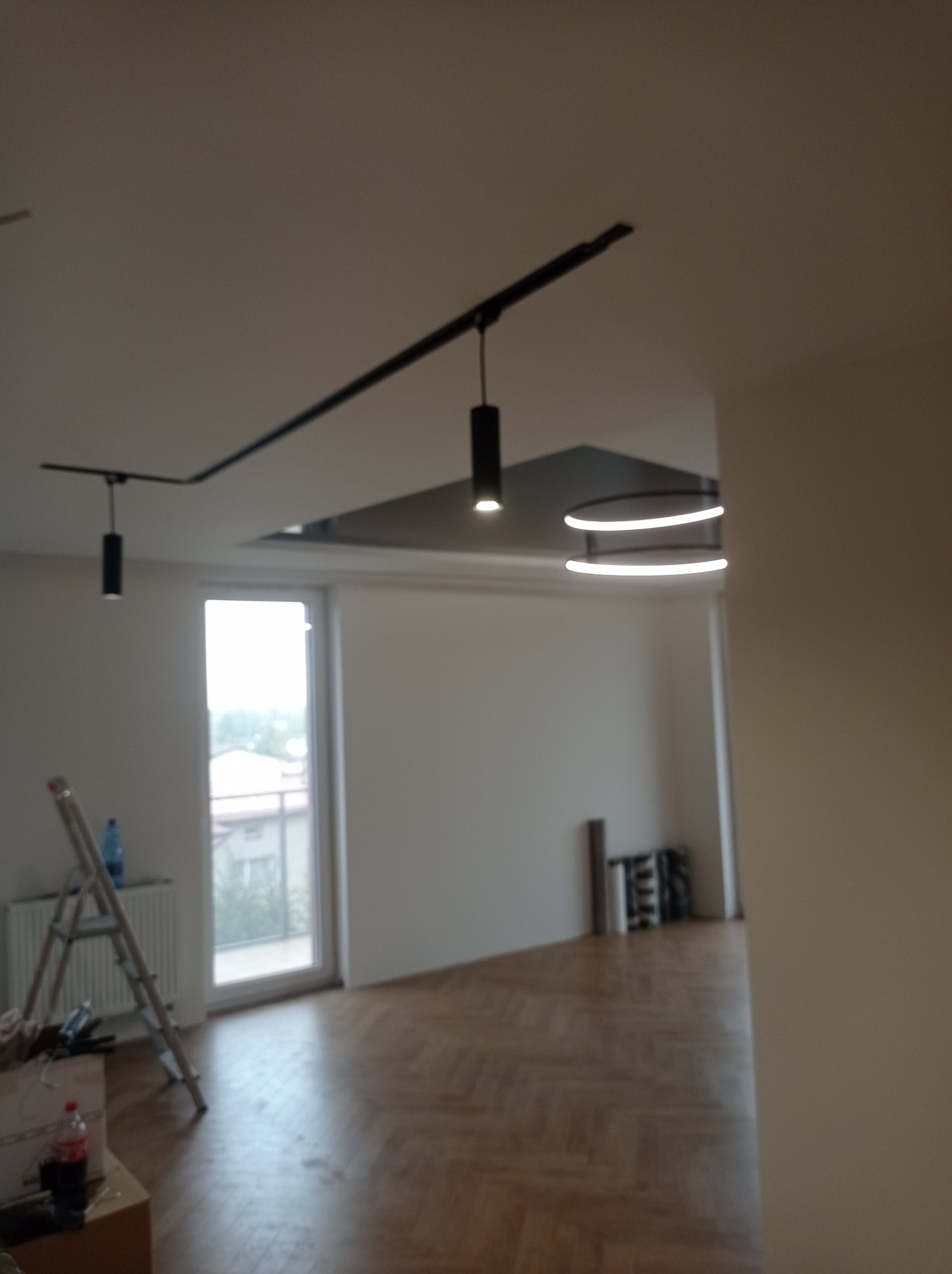 Wnętrze w trakcie prac: drewniana podłoga w jodełkę, białe ściany, zamontowane nowoczesne oświetlenie sufitowe, drabina oparta o ścianę przy oknie.