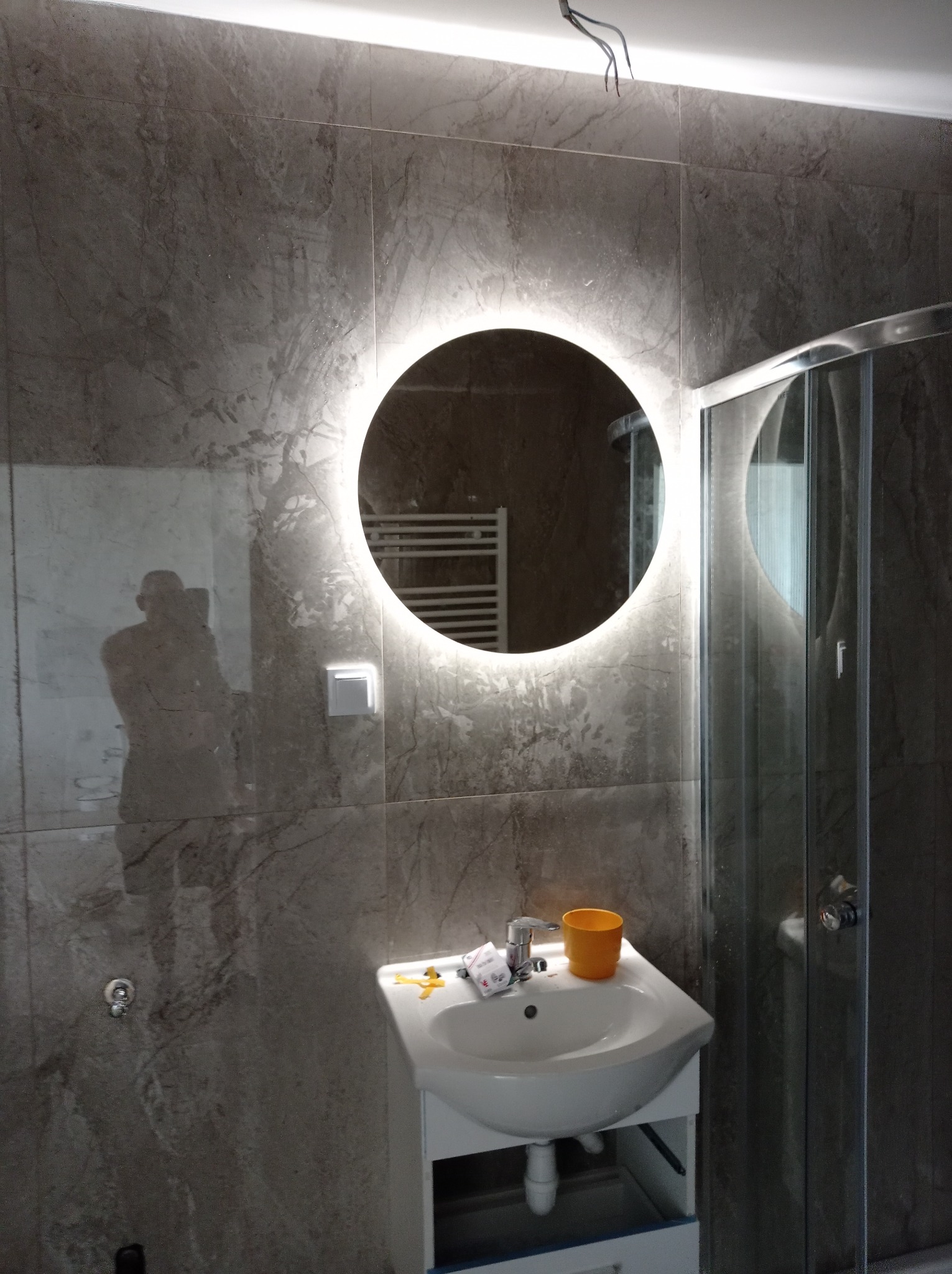 Nowoczesna łazienka z umywalką, okrągłym lustrem z podświetleniem LED i kabiną prysznicową, wykończona płytkami imitującymi marmur, z widocznym odbiciem osoby robiącej zdjęcie.