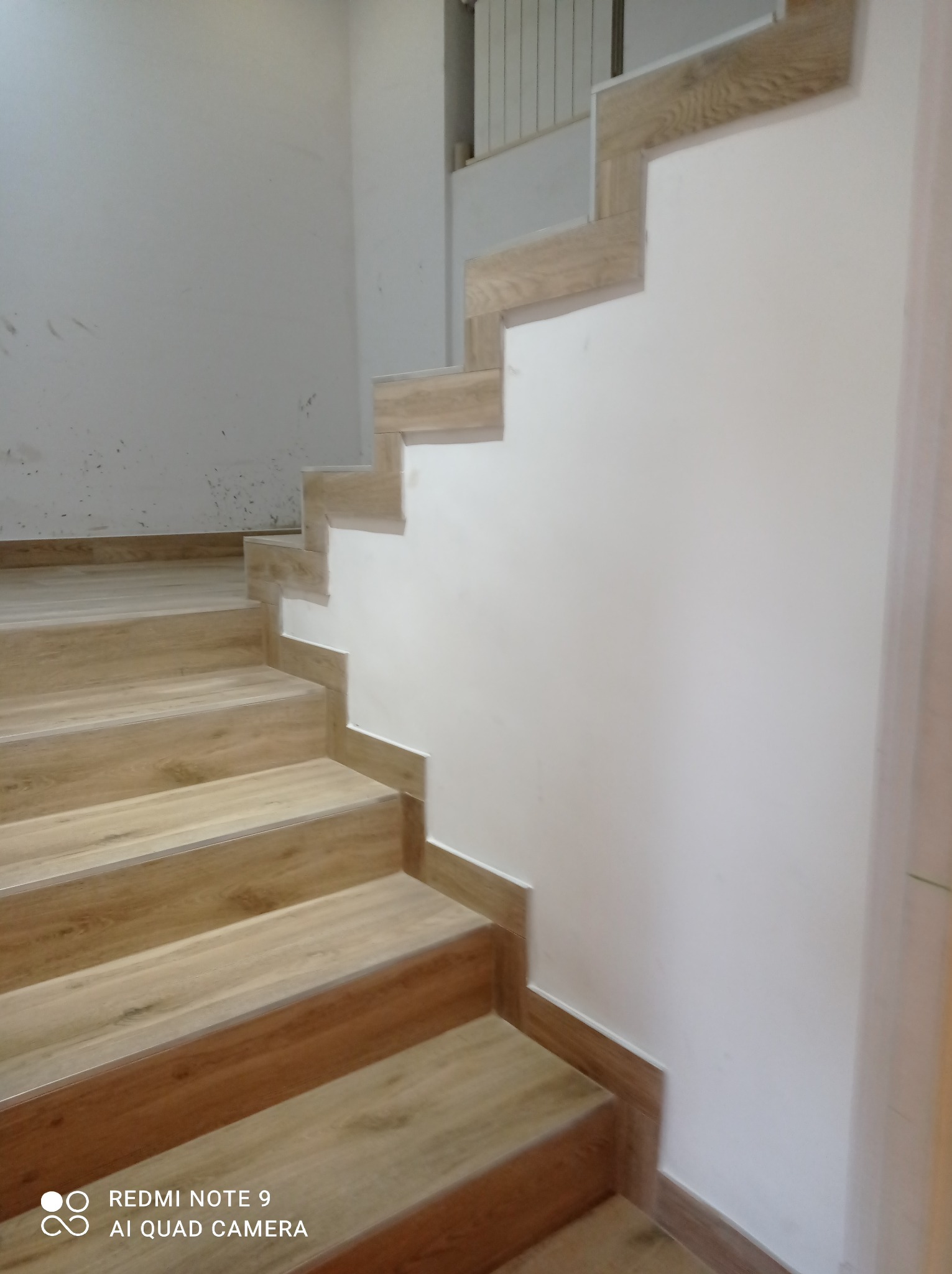 Schody wykończone panelami imitującymi drewno, z białymi ścianami, widoczne niedociągnięcia na ścianie.