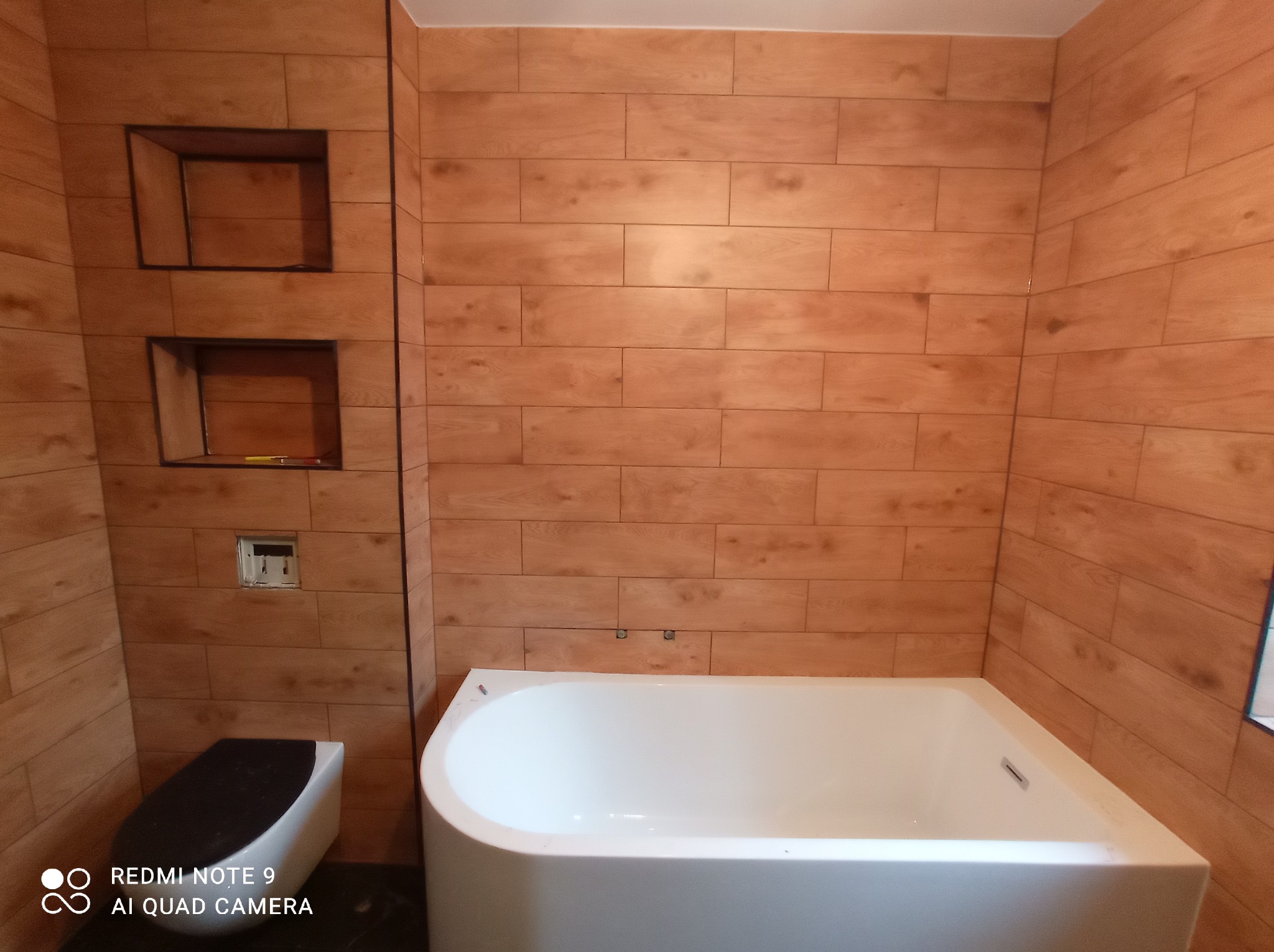 Wykończona łazienka z wanną, toaletą i ścianami wyłożonymi płytkami imitującymi drewno, z wnękami w ścianie.