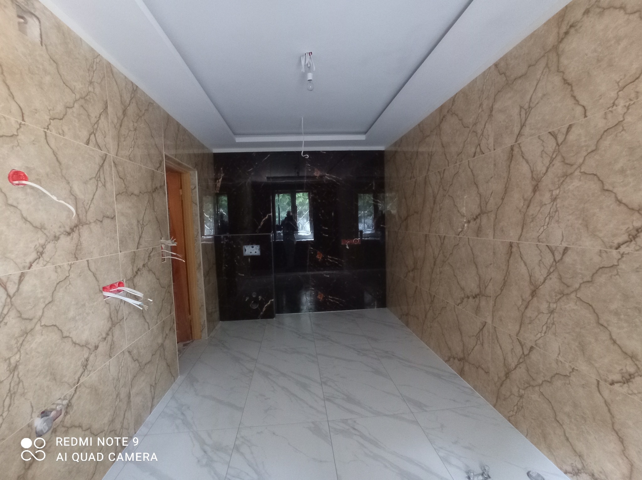 Wnętrze łazienki w trakcie wykańczania, widoczne marmurowe płytki na ścianach i podłodze, wystające kable elektryczne i surowa żarówka na suficie.