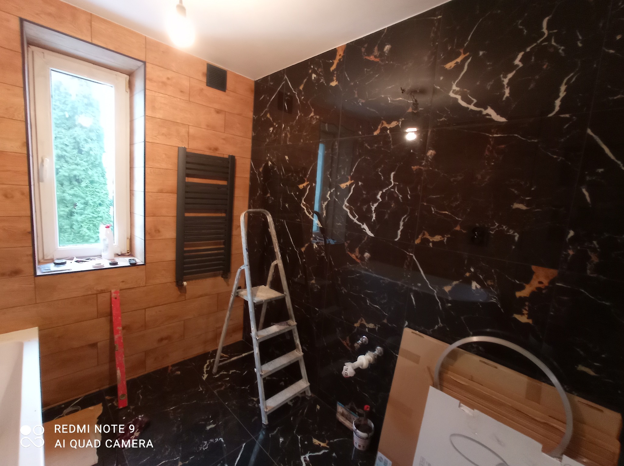 Remont łazienki w trakcie: brązowe płytki imitujące drewno na ścianie z oknem, czarne płytki z białymi żyłami na drugiej ścianie, drabina aluminiowa, grzejnik drabinkowy, poziomica, elementy...
