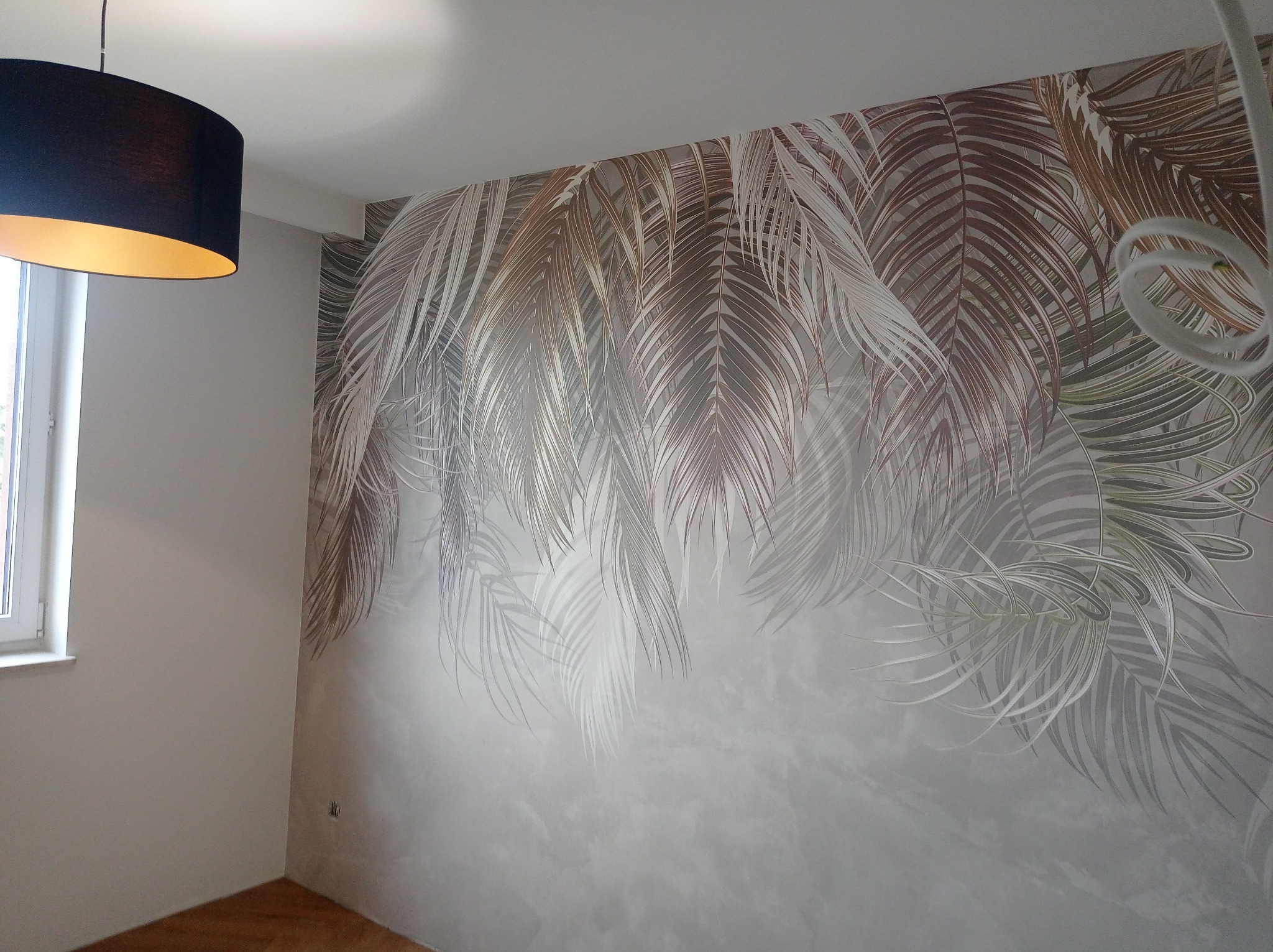Ściana w pokoju z dekoracyjną tapetą w motywie liści palmowych, oświetlona lampą z abażurem. Widoczny fragment okna i parkietu. Wykończenie wnętrza w odcieniach szarości i brązu.
