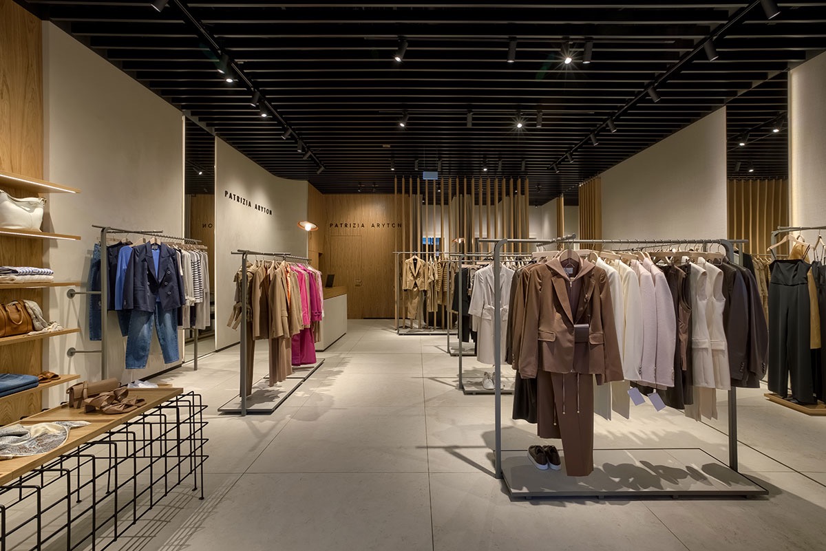 Wnętrze butiku Patrizia Aryton z ekspozycją ubrań na wieszakach i półkach, widok na minimalistyczny wystrój i oświetlenie punktowe.