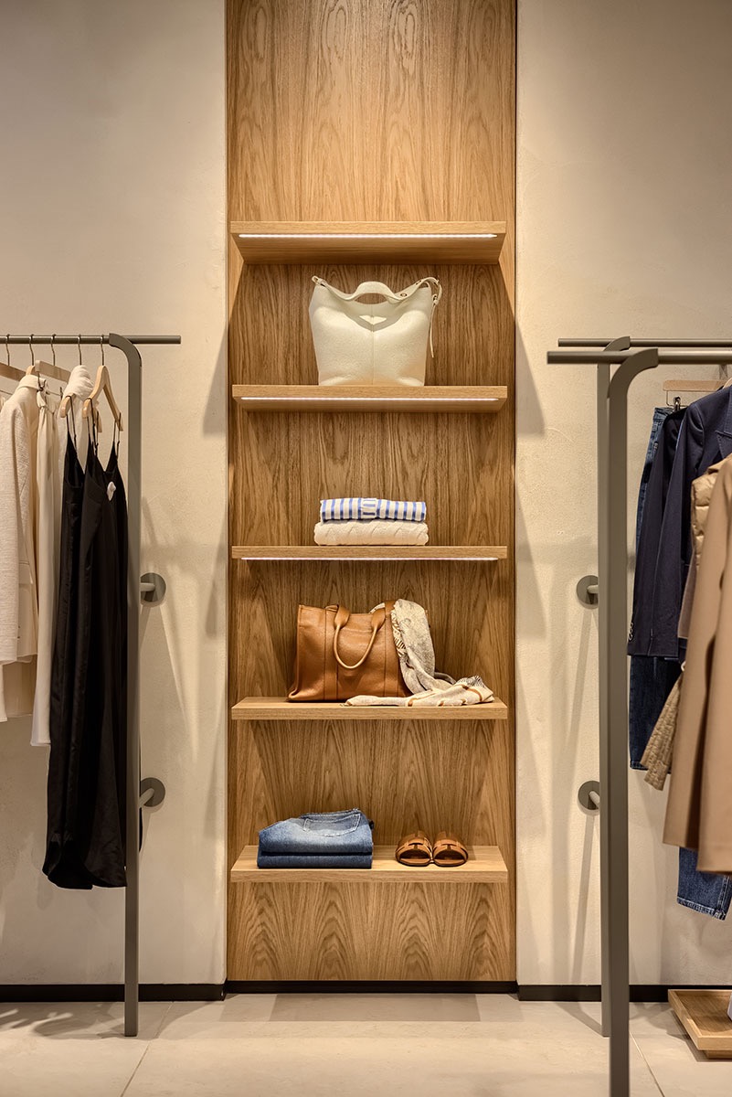 Wnętrze butiku: drewniany regał z podświetlonymi półkami, na których ułożone są torebki, ubrania i buty; po bokach wieszaki z odzieżą.