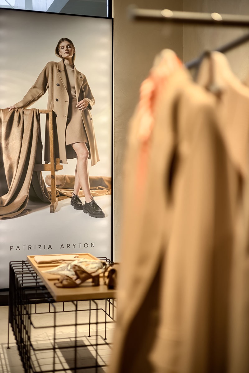 Wnętrze butiku Patrizia Aryton w Zabrzu: plakat reklamowy z modelką w beżowym płaszczu i sukience, oraz wieszaki z ubraniami w tle.