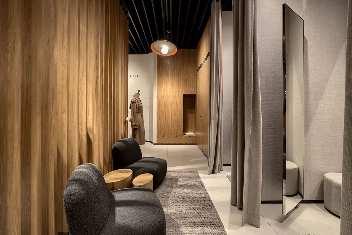 Wnętrze butiku Patrizia Aryton z drewnianymi panelami ściennymi, szarą wykładziną dywanową, dwoma ciemnymi fotelami i stolikami kawowymi z drewnianych pniaków, oraz przymierzalniami z zasłonami.