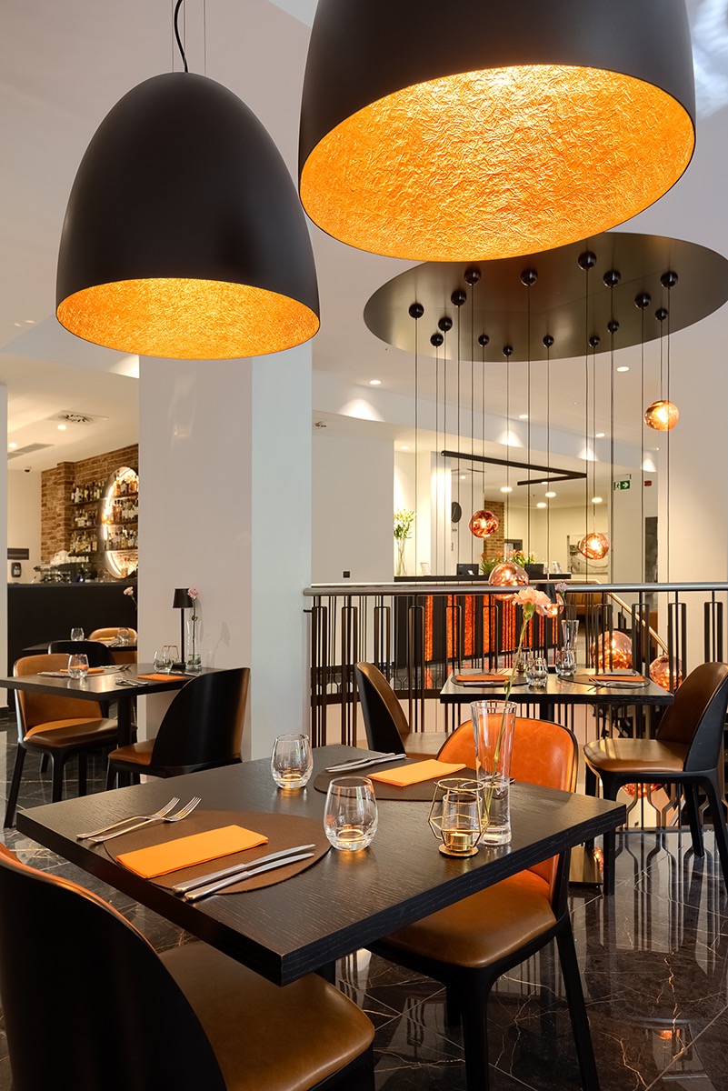 Eleganckie wnętrze restauracji hotelowej z czarnymi stołami, pomarańczowymi akcentami i designerskimi lampami wiszącymi o złotej poświacie.