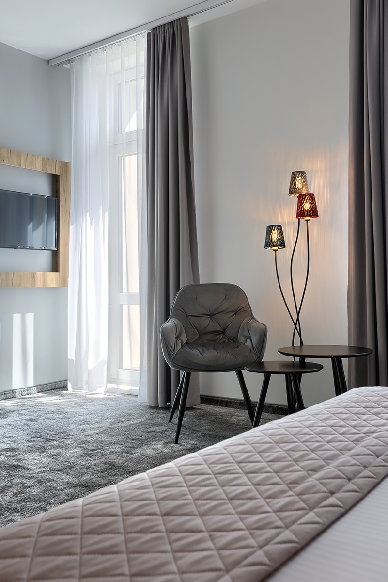 Pokój hotelowy z szarym fotelem, lampą z trzema abażurami w kolorach grafitowym, czerwonym i niebieskim oraz łóżkiem z beżową narzutą w geometryczne wzory.