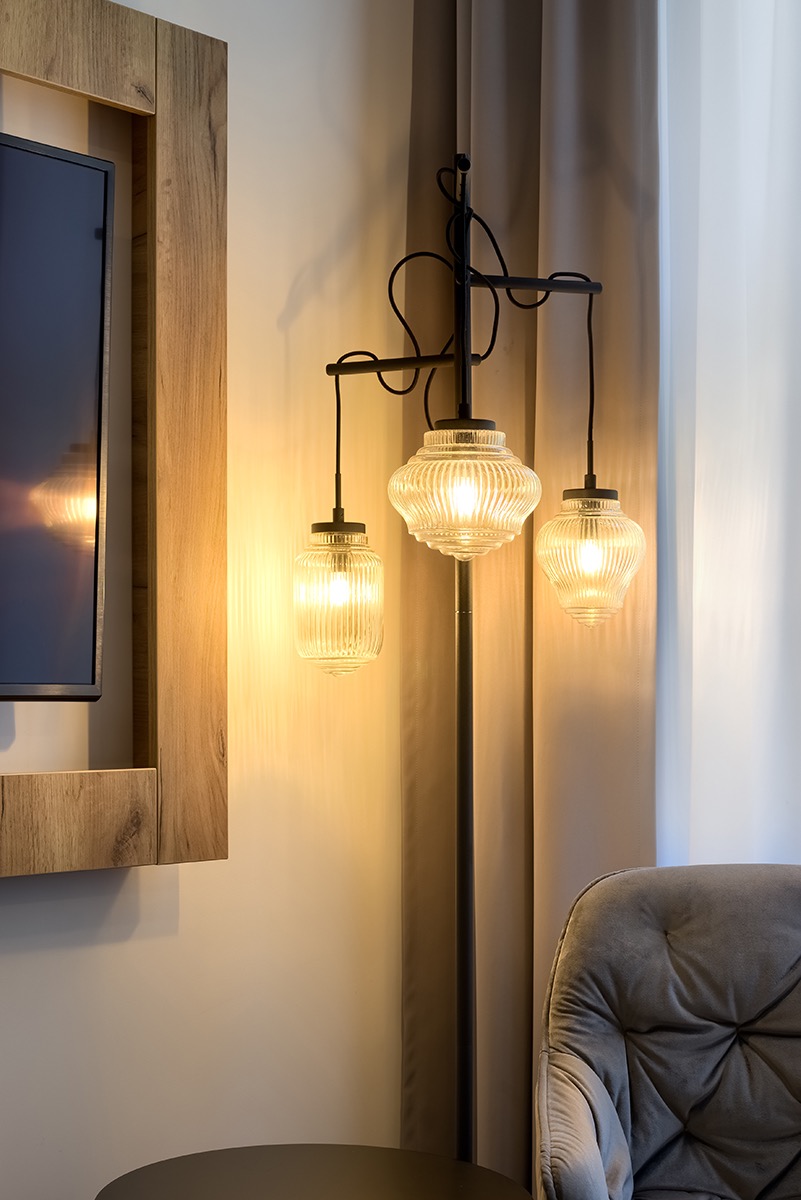 Oświetlona, trójramienna lampa podłogowa z ryflowanymi szklanymi kloszami w hotelowym wnętrzu, obok telewizor w drewnianej ramie i szary fotel.