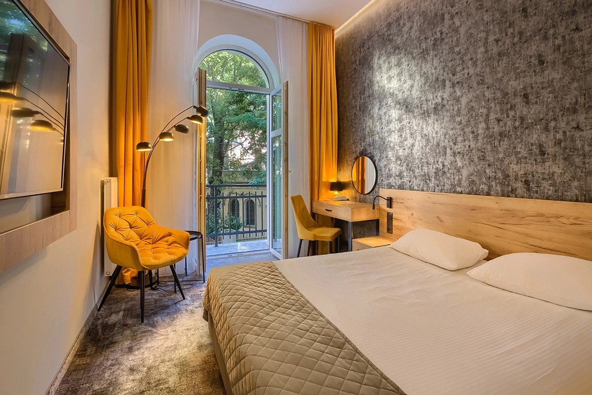 Pokój hotelowy z balkonem: żółty fotel, lampa łukowa, biurko z okrągłym lustrem, łóżko z pikowaną narzutą, ściana z efektem betonu.