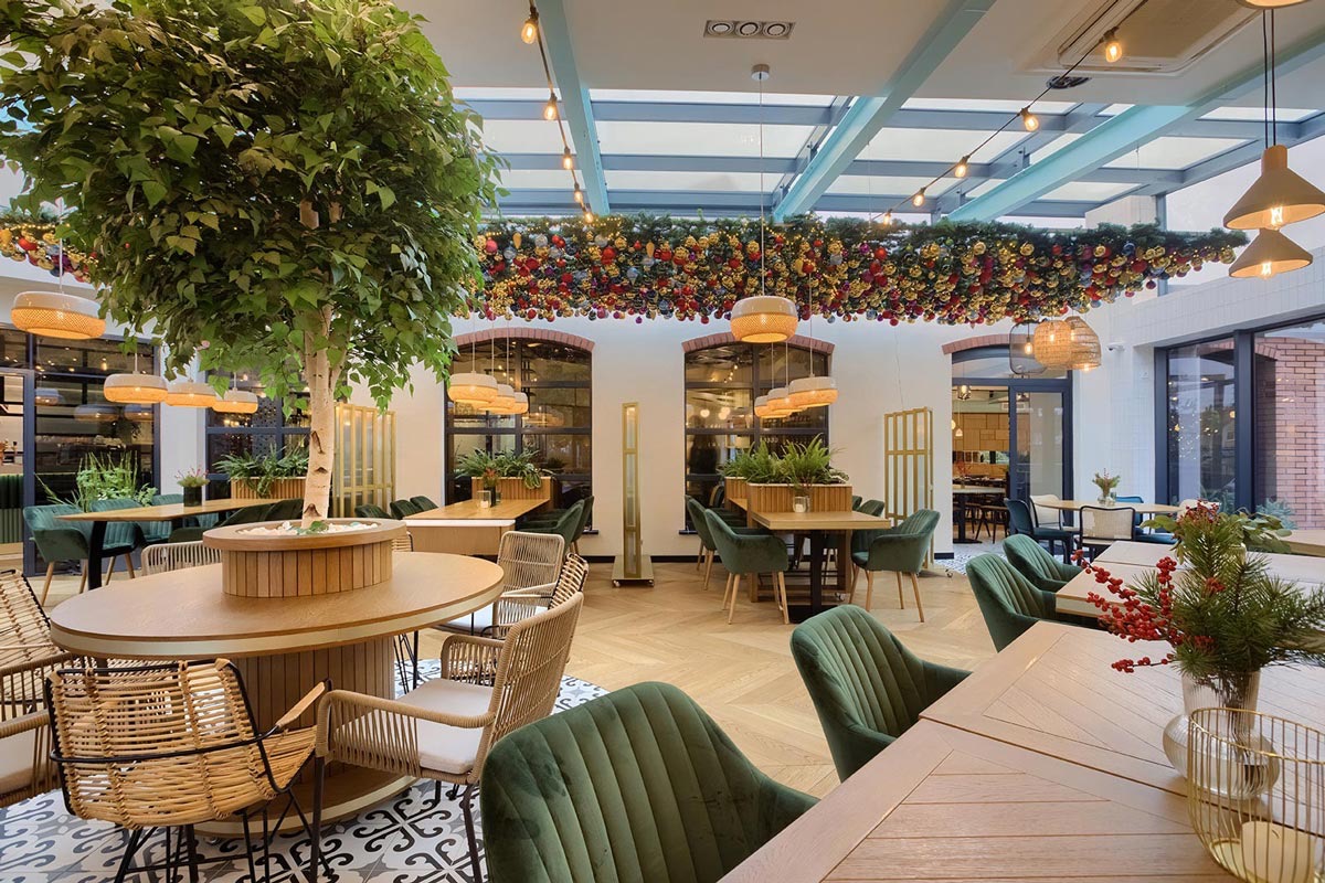 Wnętrze restauracji z dużą ilością zieleni, w tym drzewo w centralnej części sali, dekoracje świąteczne pod szklanym sufitem, drewniane stoły i krzesła z zielonym obiciem.