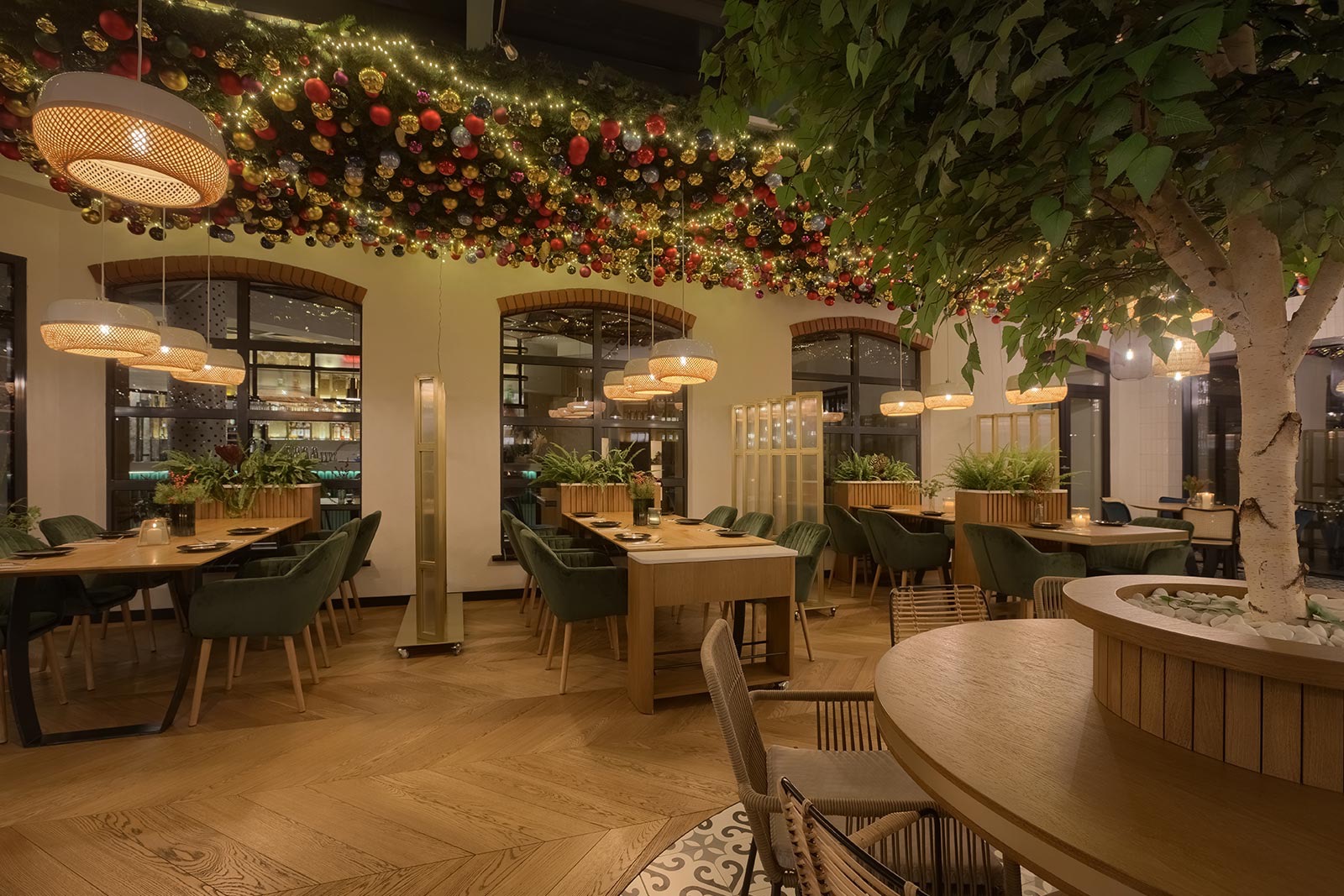 Wnętrze restauracji z dekoracją świąteczną na suficie, drewnianymi stołami i zielonymi krzesłami, oświetlone lampami z abażurami z plecionki, z drzewem w donicy po prawej stronie.