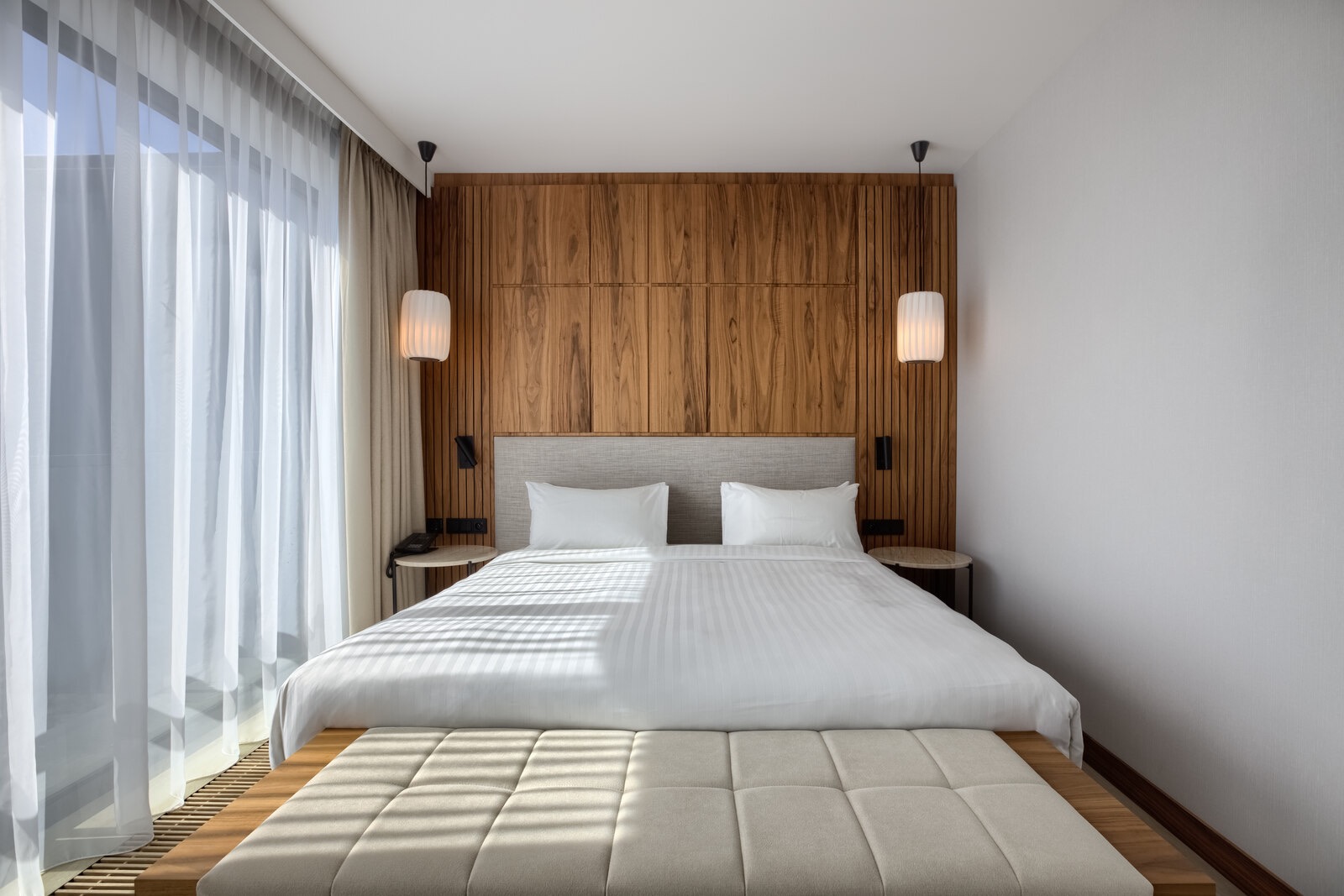 Nowoczesna sypialnia w hotelu z drewnianą ścianą za łóżkiem, lampkami nocnymi i puszystą ławeczką u podnóża.