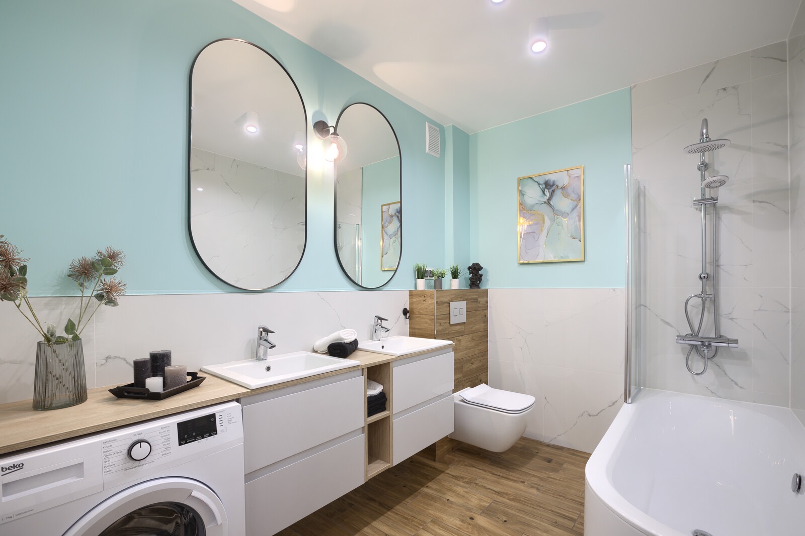 Jasna łazienka z drewnianą podłogą, umywalkami nablatowymi, pralką i wanną z prysznicem, akcentowana miętową ścianą i owalnymi lustrami.