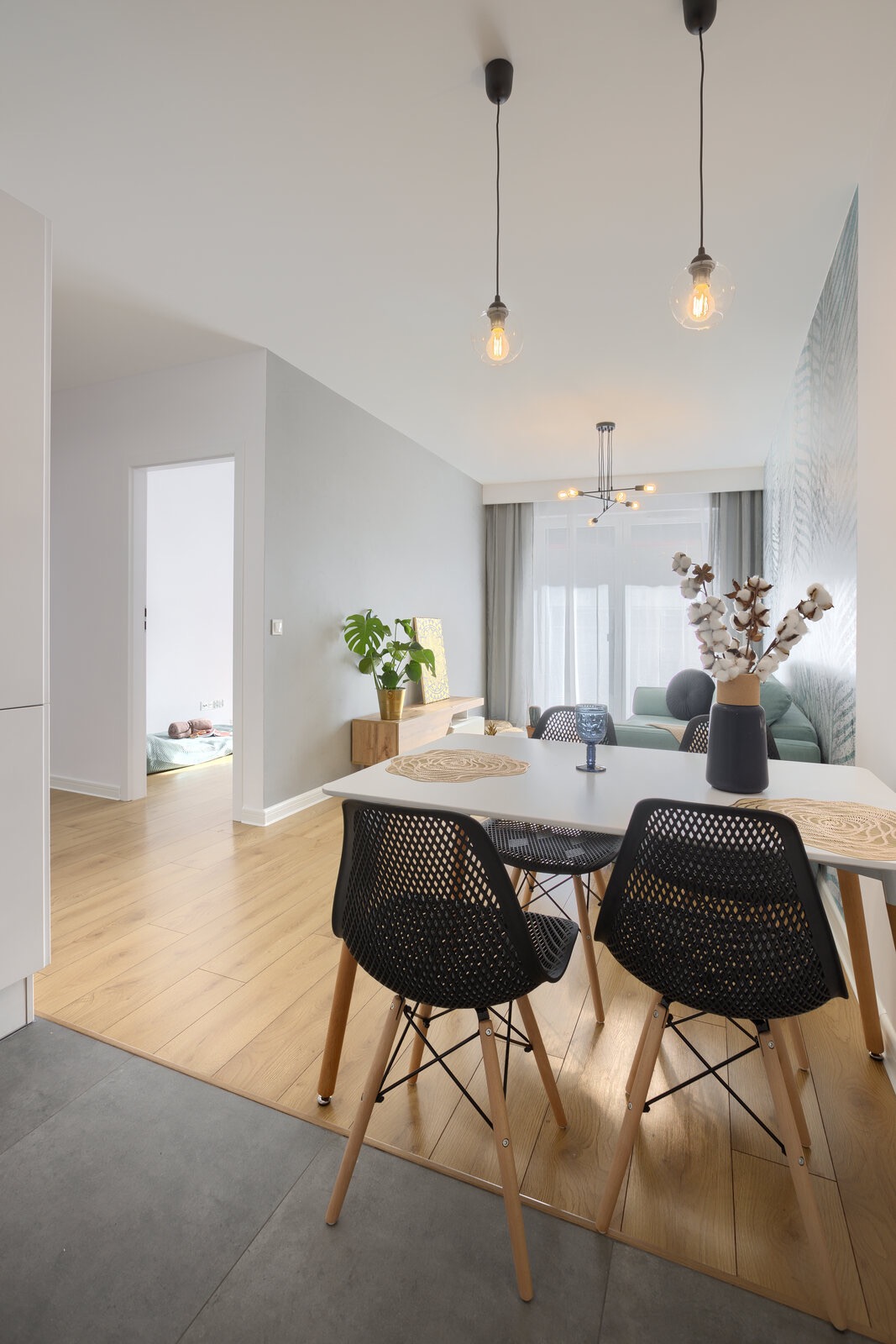 Jasne wnętrze apartamentu z białym stołem, czarnymi krzesłami z drewnianymi nogami, dekoracją z bawełny w wazonie i widokiem na pokój dziecięcy z leżącym dzieckiem.