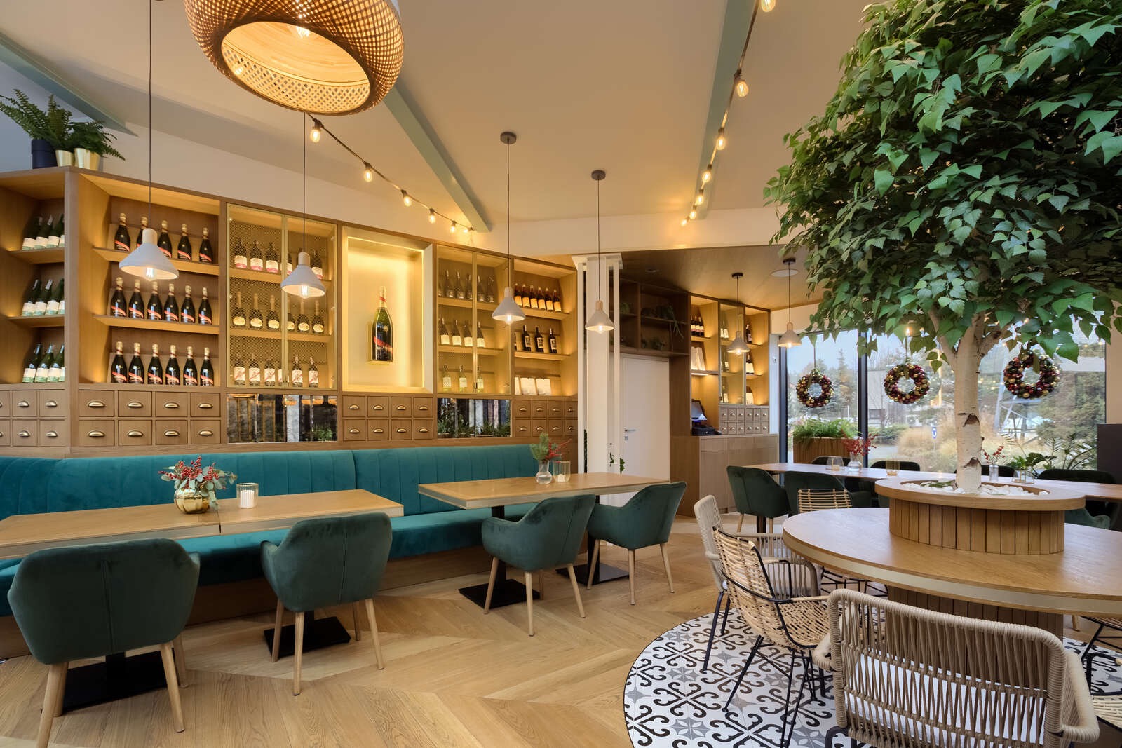 Wnętrze restauracji z drewnianą podłogą w jodełkę, turkusową sofą wzdłuż ściany z podświetlonymi półkami pełnymi butelek wina, okrągłym dywanem w stylu vintage i dekoracyjnym drzewem przy oknie.