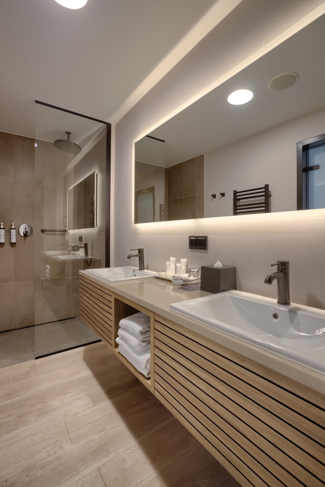 Nowoczesna łazienka z dwoma umywalkami, podświetlanym lustrem, kabiną prysznicową typu walk-in i drewnianymi szafkami z ręcznikami.