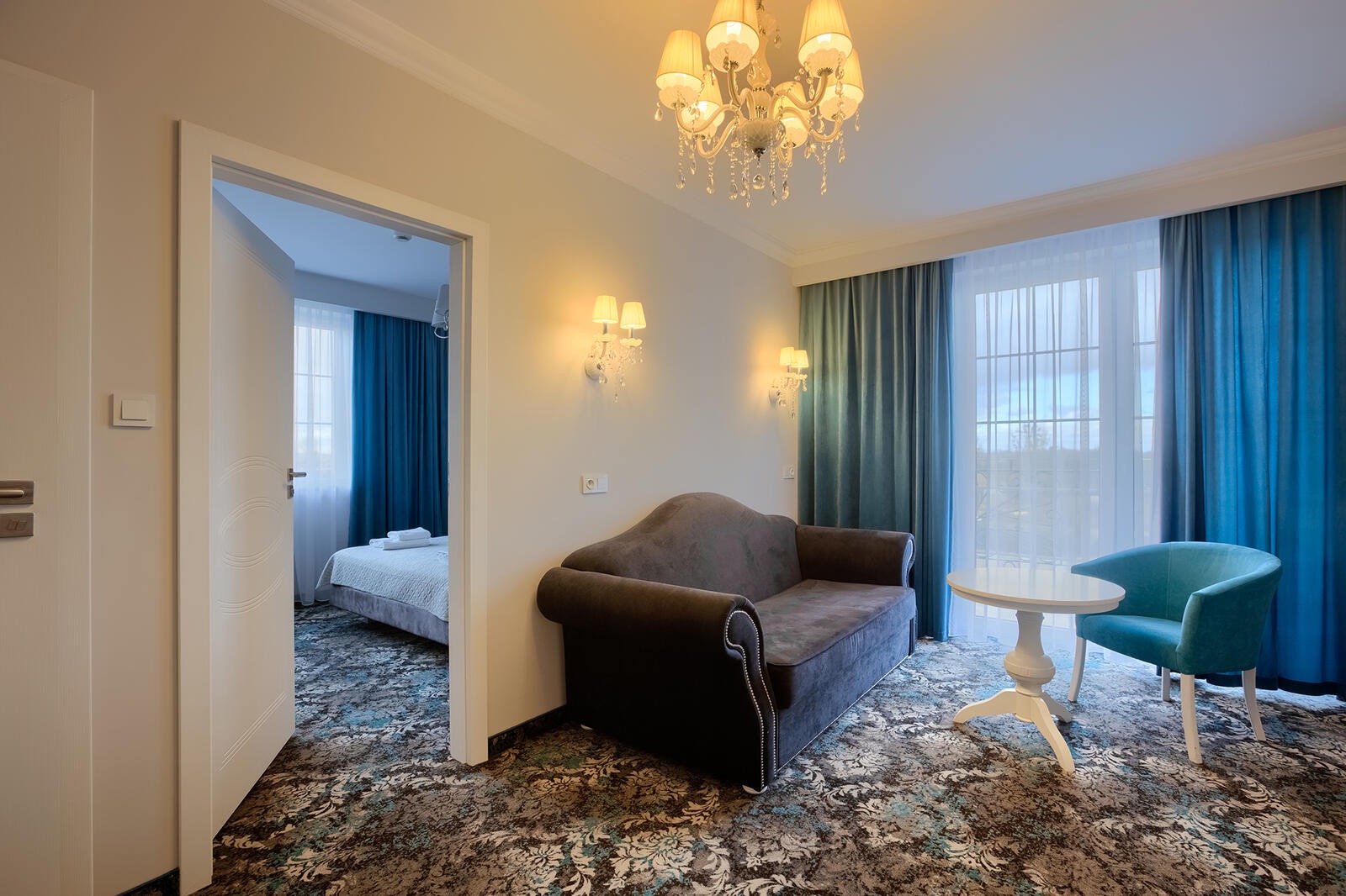 Wnętrze pokoju hotelowego z szarą sofą, turkusowym fotelem i widokiem na sypialnię z łóżkiem przez otwarte drzwi.