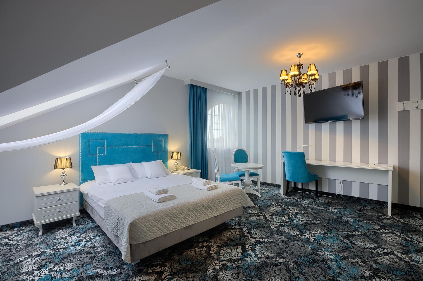 Pokój hotelowy z turkusowym zagłówkiem łóżka, białymi ręcznikami, lampkami nocnymi, stolikiem z krzesłami i biurkiem pod telewizorem na tle ściany w szaro-białe pasy.