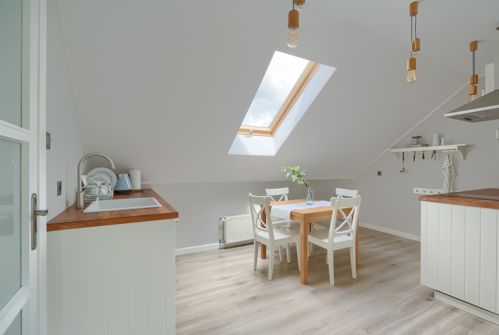Jasna kuchnia na poddaszu z drewnianym stołem i białymi krzesłami, oświetlona przez okno dachowe i wiszące lampy z żarówkami Edisona.