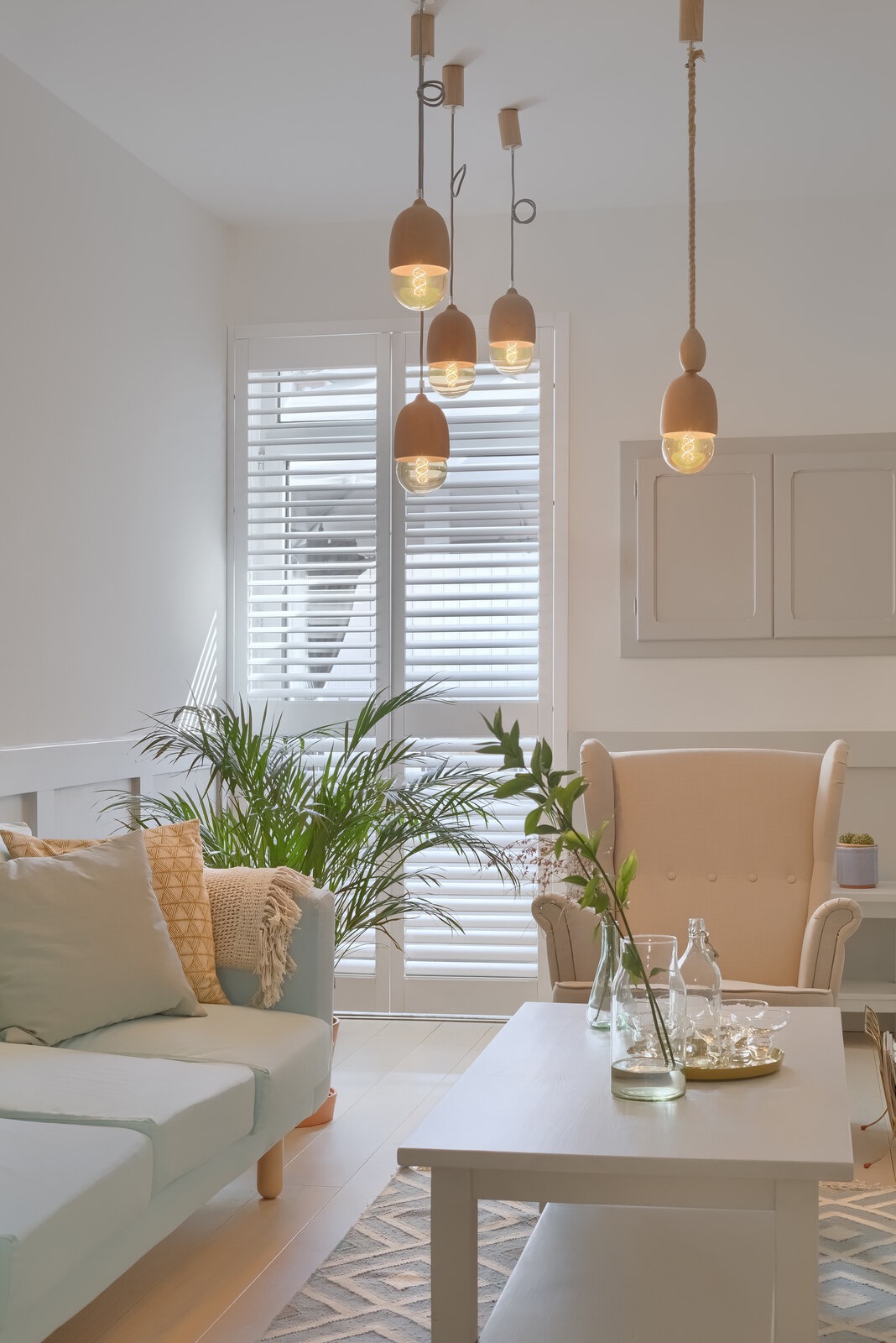 Jasne wnętrze salonu z pastelową sofą, drewnianym stolikiem kawowym, beżowym fotelem i oryginalnymi lampami wiszącymi z drewnianymi elementami.