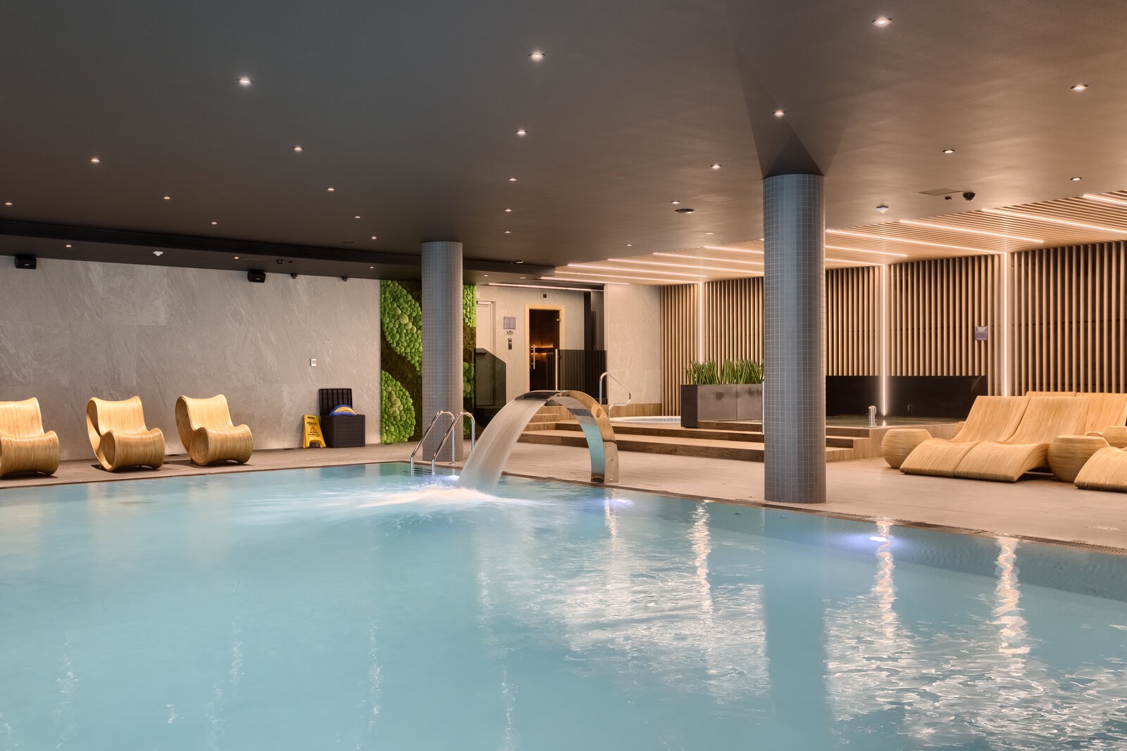 Wnętrze hotelowego basenu z wodospadem, drewnianymi leżakami i krzesłami o organicznym kształcie, oświetlone punktowymi lampami.