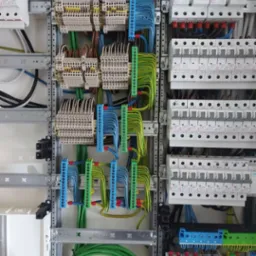 Rozdzielnica elektryczna z licznymi przewodami w kolorach: białym, zielonym, niebieskim i brązowym, podłączonymi do listew zaciskowych i zabezpieczeń, zamontowana w metalowej obudowie.