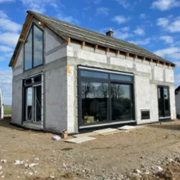 Surowy budynek w trakcie budowy z zamontowanymi oknami i drzwiami w ciemnych ramach, widoczny częściowo pokryty dach i białe bloczki konstrukcyjne, teren wokół budynku niezagospodarowany.