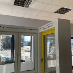 Ekspozycja w salonie: dwuskrzydłowe okno z profili PCV w kolorze białym oraz drzwi balkonowe z żółtą ramą, widoczne przekroje profili okiennych na parapecie, w tle ulica z przejściem dla pieszych.