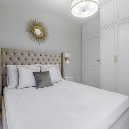 POWER-LAK - Jasna sypialnia z łóżkiem z tapicerowanym wezgłowiem, lustrem w kształcie słońca nad nim i dużą białą szafą z frezowanymi drzwiami.