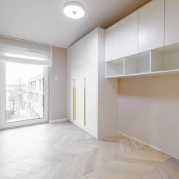 POWER-LAK - Jasne wnętrze z białą szafą wnękową z złotymi uchwytami, drewnianą podłogą ułożoną w jodełkę i oknem z roletą rzymską.