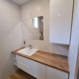 Nowoczesna łazienka w Kłocku z białymi szafkami pod umywalką z drewnianym blatem i wiszącą szafką nad umywalką, widok na lustro i wzorzyste płytki na ścianie.