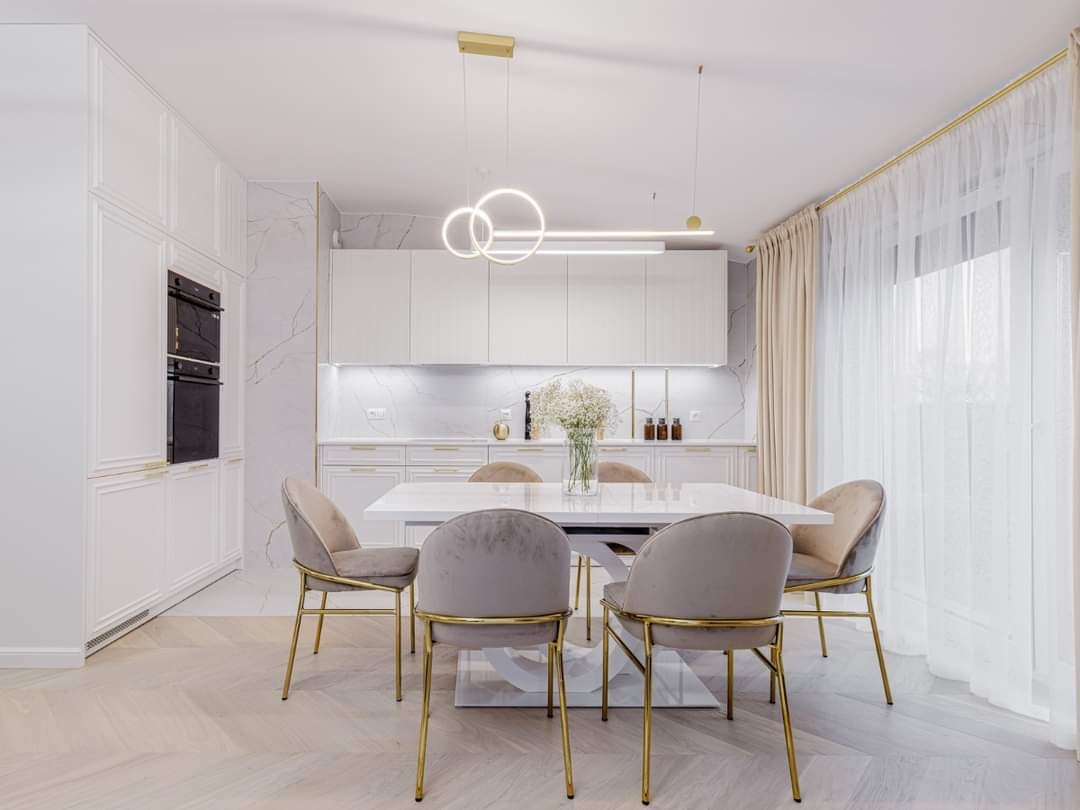 Jasna kuchnia z białymi frontami i marmurowym backsplashiem, stół z krzesłami w stylu glamour, nowoczesne oświetlenie wiszące, parkiet ułożony w jodełkę.