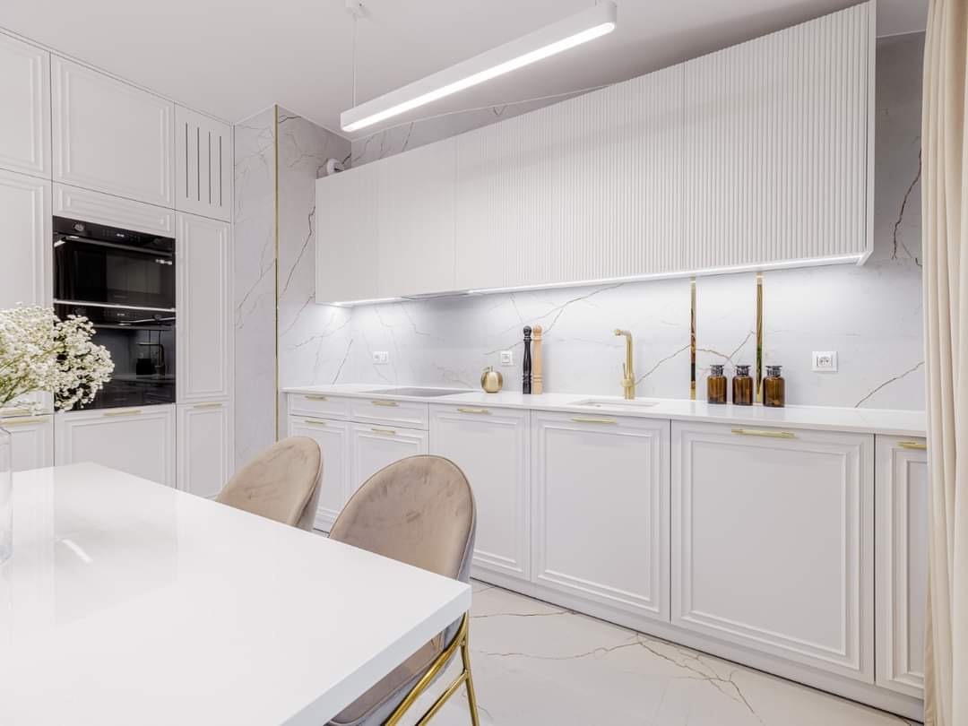 Elegancka, biała kuchnia z marmurowym blatem i złotymi akcentami, oświetlona lampą LED, z widocznym stołem i krzesłami w stylu glamour.