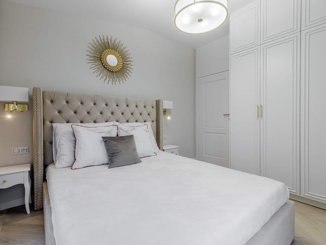 Jasna sypialnia z łóżkiem z tapicerowanym wezgłowiem, lustrem w kształcie słońca nad nim i dużą białą szafą z frezowanymi drzwiami.