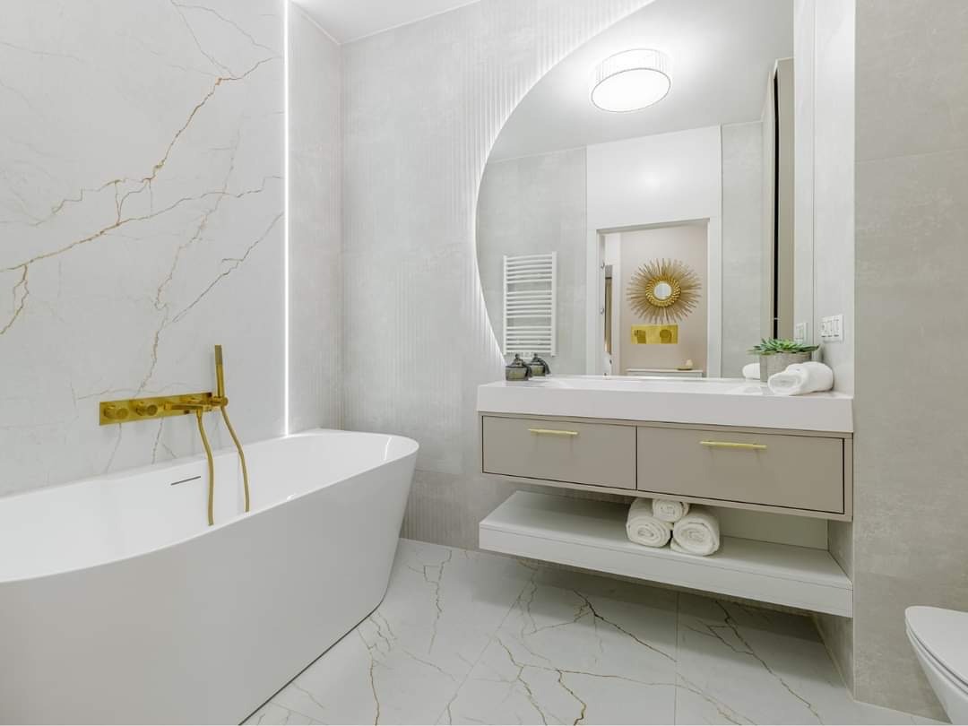 Nowoczesna łazienka z białą wanną wolnostojącą, złotą armaturą, umywalką z szafką w odcieniach szarości i okrągłym lustrem z podświetleniem LED.