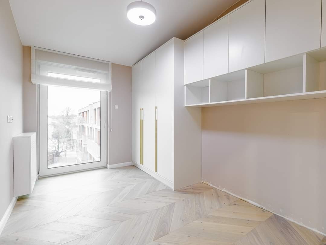 Jasne wnętrze z białą szafą wnękową z złotymi uchwytami, drewnianą podłogą ułożoną w jodełkę i oknem z roletą rzymską.