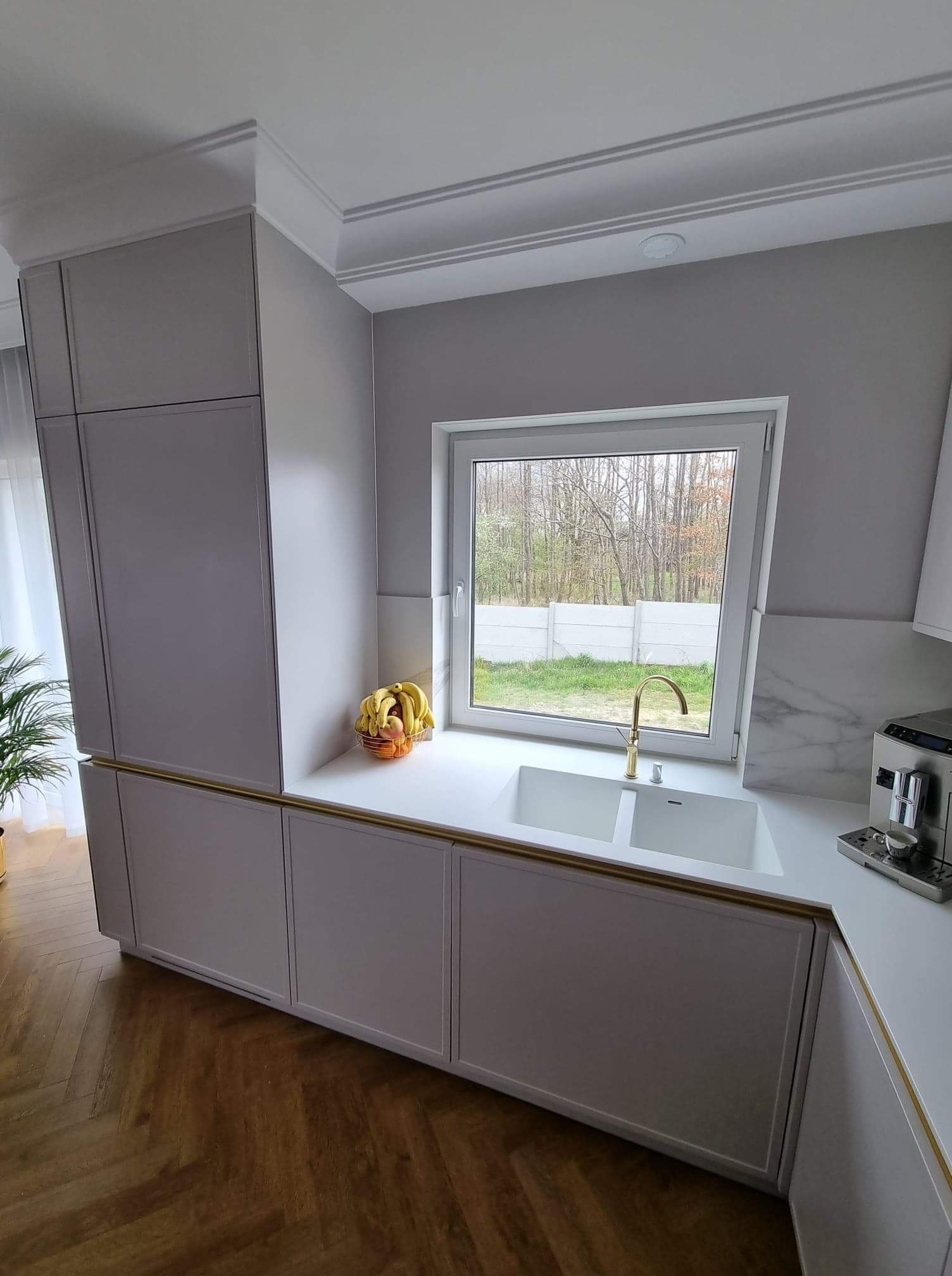 Nowoczesna kuchnia w odcieniach szarości z białym blatem, złotą armaturą i drewnianą podłogą w jodełkę, widok na ogród przez okno.