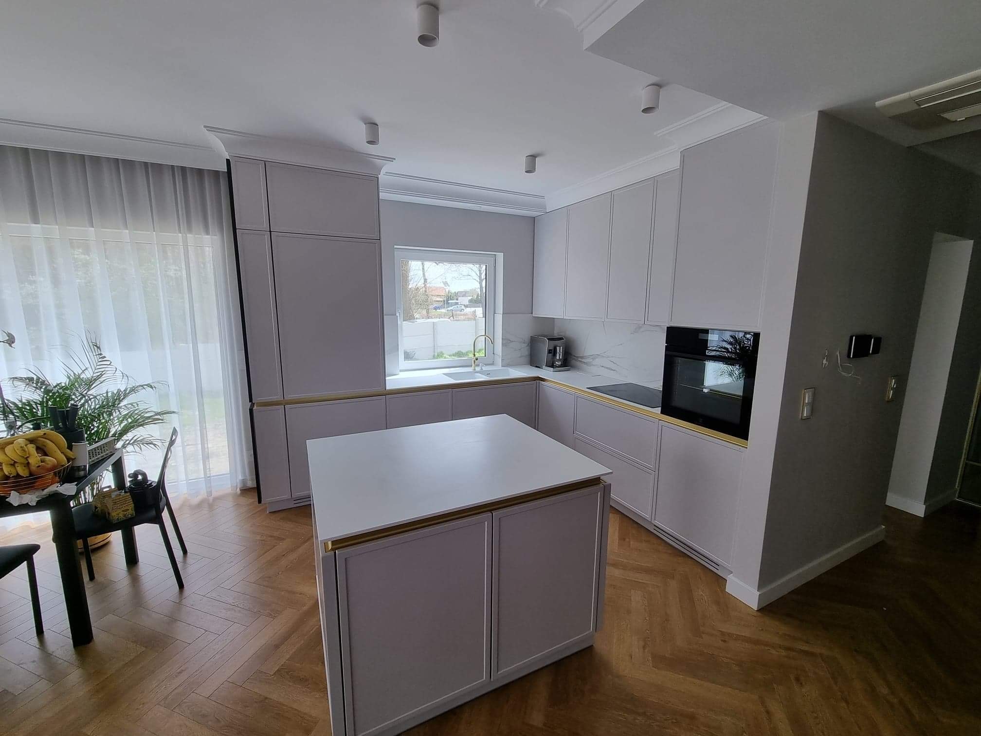 Jasna kuchnia z wyspą, fronty szafek z delikatnym frezem i złotymi akcentami, parkiet w jodełkę, widok na okno i stół z owocami.